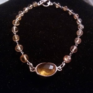 K&ouml;nnte beinhalten: Ein silbernes Armband mit einem gro&szlig;en, ovalen, bernsteinfarbenen Edelstein in der Mitte. Das Armband besteht aus kleinen, runden, bernsteinfarbenen Perlen.