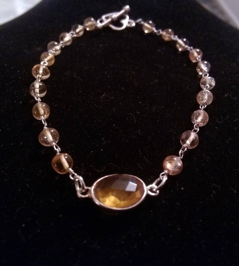 K&ouml;nnte beinhalten: Ein silbernes Armband mit einem gro&szlig;en, ovalen, bernsteinfarbenen Edelstein in der Mitte. Das Armband besteht aus kleinen, runden, bernsteinfarbenen Perlen.