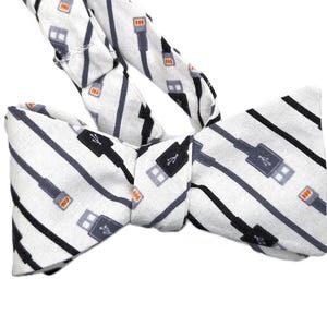 May include: A white bow tie featuring a repeating pattern of black, grey, and orange USB cable designs. The fabric bow tie has a classic shape with adjustable straps for a comfortable fit.
