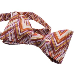 Può includere: Un papillon fantasia con un design geometrico nei toni del viola, arancione e oro su sfondo crema. Il papillon è annodato e pronto da indossare, mostrando un accessorio elegante.