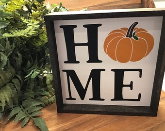May include: A wooden sign with a black frame and a white background. The sign says "HOME" with a pumpkin in the middle. The pumpkin is orange with a green stem.