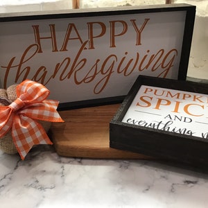 May include: Two framed signs with fall-themed messages. The larger sign says "HAPPY thanksgiving" in orange script on a white background. The smaller sign says "PUMPKIN SPICE AND everything nice" in orange text on a white background. A small burlap pumpkin with an orange and white checkered bow sits in front of the signs.