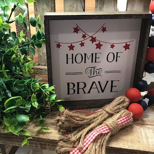 Może przedstawiać: Drewniany znak z napisem "Home of the Brave" czarnymi literami. Znak jest ozdobiony czerwonymi gwiazdami i czerwono-biało-niebieską girlandą z koralik&oacute;w.