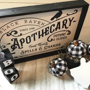 May include: Black and white sign with the text "Black Raven Apothecary - Since 1790 - Finest Quality Spells & Charms - Potions & Elixirs". The sign is framed in black wood and has a black raven silhouette. There are three black and white checkered fabric pumpkins and a white ghost figurine on a white surface.