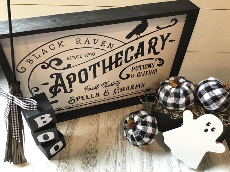 May include: Black and white sign with the text "Black Raven Apothecary - Since 1790 - Finest Quality Spells & Charms - Potions & Elixirs". The sign is framed in black wood and has a black raven silhouette. There are three black and white checkered fabric pumpkins and a white ghost figurine on a white surface.