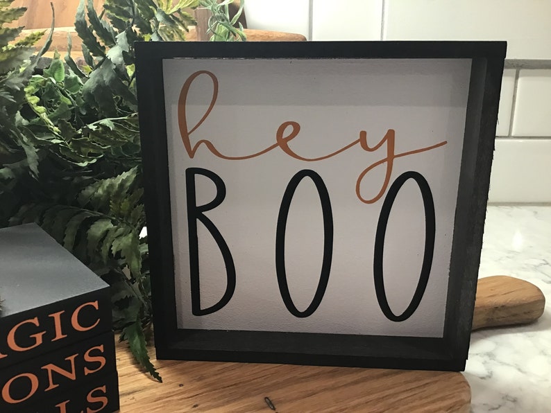 May include: A black framed wooden sign with a white background and the text "hey boo" in black and orange lettering.