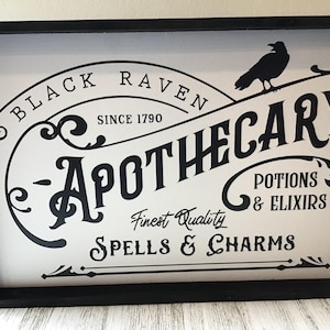 May include: Black and white sign with the text "Black Raven Apothecary - Finest Quality Spells & Charms - Potions & Elixirs" and a silhouette of a raven. The sign is framed in black.