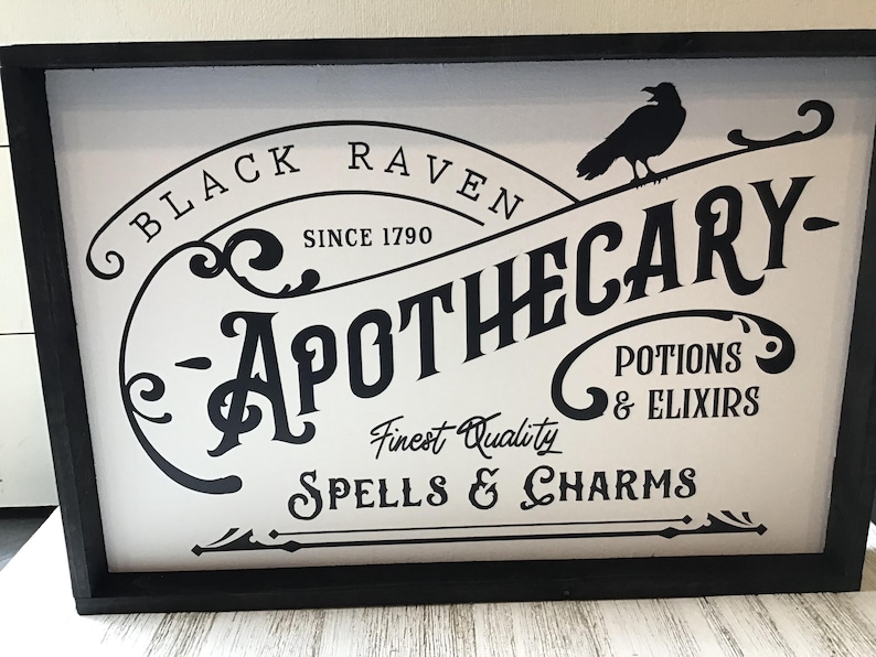 May include: Black and white sign with the text "Black Raven Apothecary - Finest Quality Spells & Charms - Potions & Elixirs" and a silhouette of a raven. The sign is framed in black.