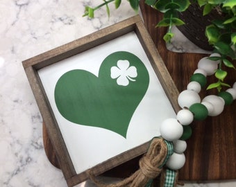May include: A wooden sign with a green heart and a white four-leaf clover in the centre. The sign is framed in a rustic brown wood frame.