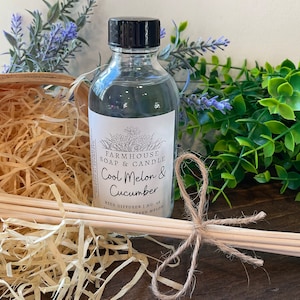 May include: A clear glass bottle with a black lid and a white label that reads "Farmhouse Soap & Candle Cool Melon & Cucumber Reed Diffuser | No. 64 Fresh Melon". The bottle is surrounded by wood-colored sticks and straw-like material.