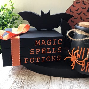 May include: Black tiered tray with orange ribbon, a black bat, a black jar with a cork lid and the text "Witches Brew" in orange, and three black blocks stacked on top of each other with the text "Magic Spells Potions" in orange.