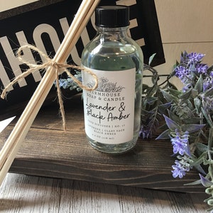 May include: A clear glass bottle with a black lid filled with a lavender and black amber scented reed diffuser. The bottle has a white label with the text "FARMHOUSE SOAP & CANDLE Lavender & Black Amber REED DIFFUSER | NO. 17 LAVENDER | CLARY SAGE BLACK AMBER 4.0 OZ (118 ml)". The bottle is sitting on a wooden surface with a bundle of wooden reeds and a sign that says "HOUSE".