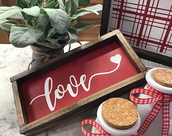 Può includere: Un cartello in legno incorniciato con sfondo rosso e lettere bianche che dice "love" con un cuore bianco e una linea bianca che si estende dal cuore.