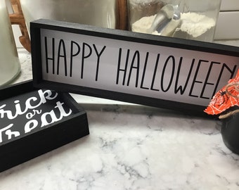 Puede incluir: Dos letreros enmarcados en negro con letras blancas. El letrero más grande dice "Happy Halloween" y el letrero más pequeño dice "Trick or Treat".