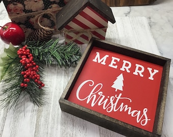 May include: A wooden framed sign with a red background and white lettering that says "Merry Christmas" with a white silhouette of a Christmas tree.