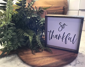 May include: A white wooden sign with black calligraphy that reads "so thankful". The sign is sitting on a wooden cutting board with a green fern plant in the background.