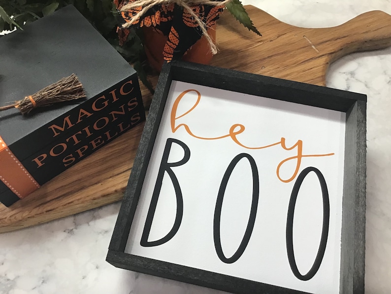May include: A black framed sign with the words "Hey Boo" in orange and black lettering on a white background.