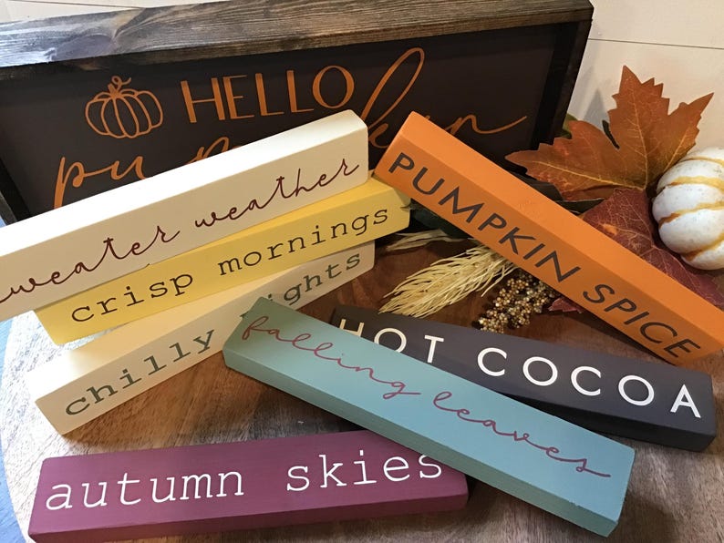 May include: A collection of six wooden signs with fall-themed text. The signs are stacked on top of each other and are arranged in a random order. The text on the signs reads: 'sweater weather', 'crisp mornings', 'chilly nights', 'pumpkin spice', 'hot cocoa', 'falling leaves', and 'autumn skies'. The signs are painted in various colors, including white, yellow, orange, brown, gray, blue, and purple.