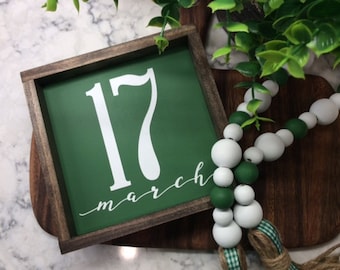 Puede incluir: Un letrero de madera verde con el número 17 y la palabra "marzo" escrita en cursiva blanca. El letrero está enmarcado en un marco de madera oscura.