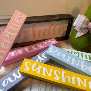 May include: A set of wooden signs with various spring-themed words, including "butterflies", "hello", "tulips", "sunshine", "rainbow", and "springtime". The signs are painted in pastel colors and have white lettering.