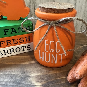 May include: Orange ceramic jar with a cork lid and twine tied around it. The jar says "Egg Hunt" in white lettering. The jar is sitting on a wooden surface with a stack of wooden blocks that say "Farm Fresh Carrots" and a couple of orange carrot decorations.