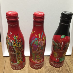 May include: Three Coca-Cola glass bottles with different designs. The first bottle has a pixelated design with the text "Pixel Art". The second bottle has a colorful design with the text "Hip Hop". The third bottle is black with the text "Zero Sugar".