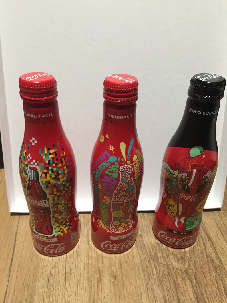 May include: Three Coca-Cola glass bottles with different designs. The first bottle has a pixelated design with the text "Pixel Art". The second bottle has a colorful design with the text "Hip Hop". The third bottle is black with the text "Zero Sugar".