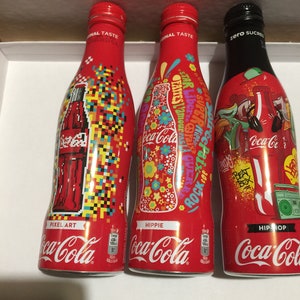 May include: Three Coca-Cola glass bottles with different designs. The bottles are red with white lettering and feature the Coca-Cola logo. The designs are Pixel Art, Hippie, and Hip-Hop.