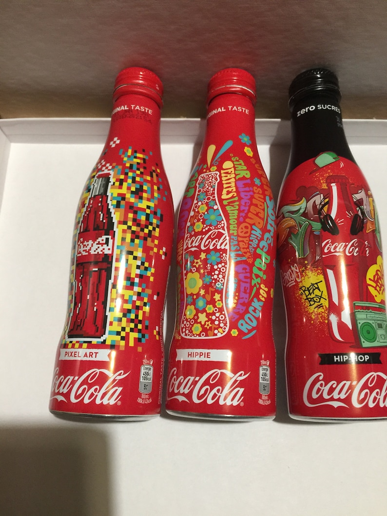 May include: Three Coca-Cola glass bottles with different designs. The bottles are red with white lettering and feature the Coca-Cola logo. The designs are Pixel Art, Hippie, and Hip-Hop.