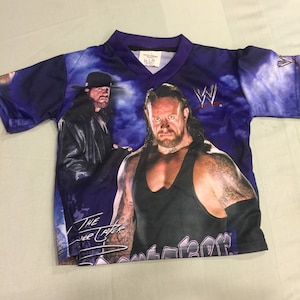 Puede incluir: Una camiseta de manga corta de color morado, negro y blanco con un estampado gráfico de dos luchadores, The Undertaker y The Big Show. La camiseta tiene el logotipo de la WWE y el texto "The Undertaker".