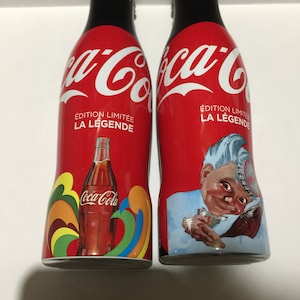 May include: Two red Coca-Cola bottles with black caps. The bottles are labeled "&Eacute;dition Limit&eacute;e La L&eacute;gende" and feature a Coca-Cola bottle and a cartoon character.