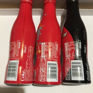 May include: Three 250ml aluminum cans of Coca-Cola. The cans are red, black, and red with a colorful design. The cans have the Coca-Cola logo and the text "Pensez au tri!" on the back.