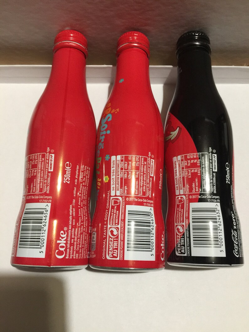 May include: Three 250ml aluminum cans of Coca-Cola. The cans are red, black, and red with a colorful design. The cans have the Coca-Cola logo and the text "Pensez au tri!" on the back.