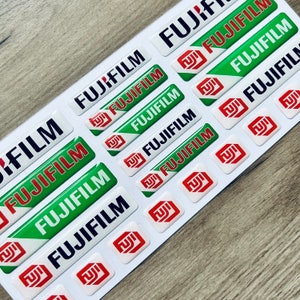Op de afbeelding: Een verzameling groene, witte en rode stickers met de tekst "FUJIFILM" erop gedrukt. De stickers zijn in een patroon op een houten oppervlak gerangschikt.
