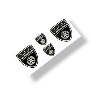 Könnte beinhalten: Vier schwarze und silberne Karmann-Aufkleber in Schildform. Jeder Aufkleber trägt das Wort "Karmann" über einem stilisierten Raddesign. Die Aufkleber befinden sich auf weißem Hintergrund.
