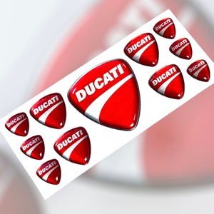 May include: A set of nine red and white Ducati motorcycle logo stickers. The stickers are shaped like a triangle with rounded corners and have a glossy finish.