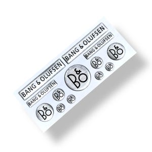 Op de afbeelding: Een vel zilverkleurige stickers met de tekst "BANG & OLUFSEN" en het "B&O" logo in zwart. De stickers hebben verschillende rechthoekige en cirkelvormige formaten, met een geborsteld metalen uiterlijk.