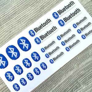May include: A sheet of white stickers with blue and black Bluetooth logos. The stickers are in various sizes and shapes, with some being rectangular and others being round. The word "Bluetooth" is printed on the rectangular stickers.