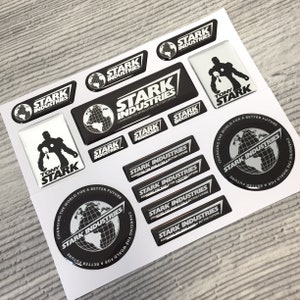 Puede incluir: Una colección de pegatinas en blanco y negro con el logotipo de Stark Industries y la frase "Cambiando el mundo para un futuro mejor". Algunas pegatinas también presentan un globo terráqueo y las palabras "Tony Stark".