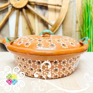 May include: An orange ceramic cooking pot with a lid, adorned with a white and black floral pattern. The pot has green accents on the handles and lid handle. The pot is sitting on a light-colored wooden surface.