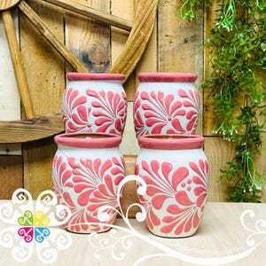 May include: Four white ceramic pots with a pink floral design. The pots have a pink rim and are stacked on top of each other. The background is a wooden wall with green plants.