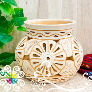 May include: Off-white ceramic vase with intricate cut-out floral and leaf designs. The vase has a wide opening and a decorative band around the top. The detailed carvings create a textured appearance, perfect for home decor.