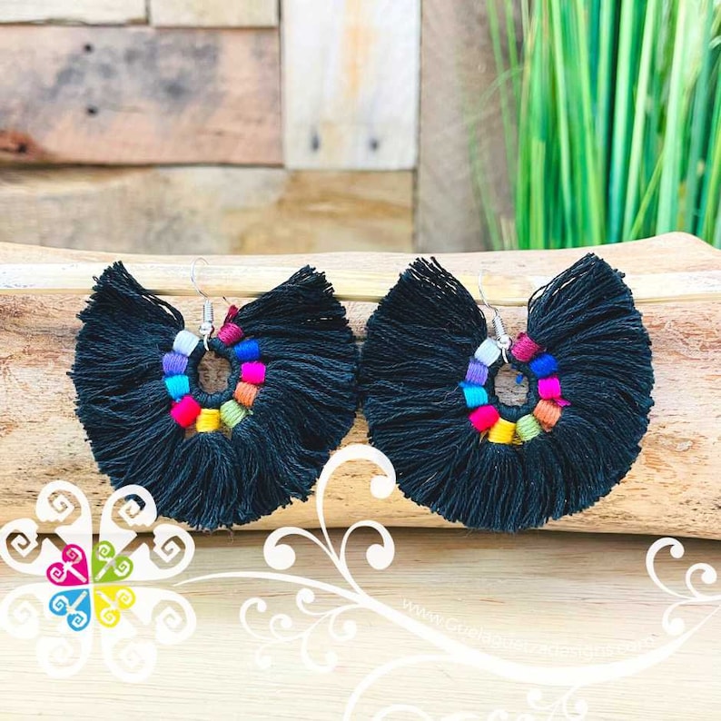 May include: A pair of black tassel earrings with a fan-like shape. The earrings feature a colorful beaded circle in the center, with a variety of colors including pink, blue, yellow, and orange. The earrings are displayed on a wooden surface.