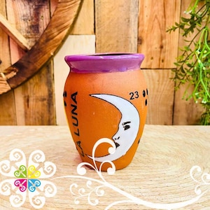 May include: Orange ceramic vase with a purple rim, featuring a white crescent moon design with a face. The vase has the text "LA LUNA" and the number "23" on it. The vase is on a woven surface.