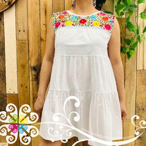 May include: White sleeveless dress with colorful floral embroidery across the neckline. The dress has a tiered skirt and is made of a lightweight fabric. The website address "www.Guelaguetzadesigns.com" is visible.