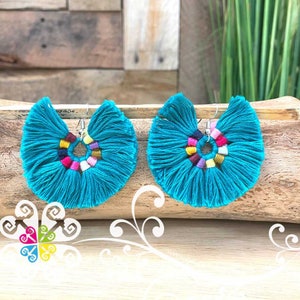 May include: A pair of turquoise tassel earrings. Each earring features a fan-shaped design with a colorful, circular accent in the center. The earrings are displayed on a wooden surface, showcasing their vibrant color and texture.