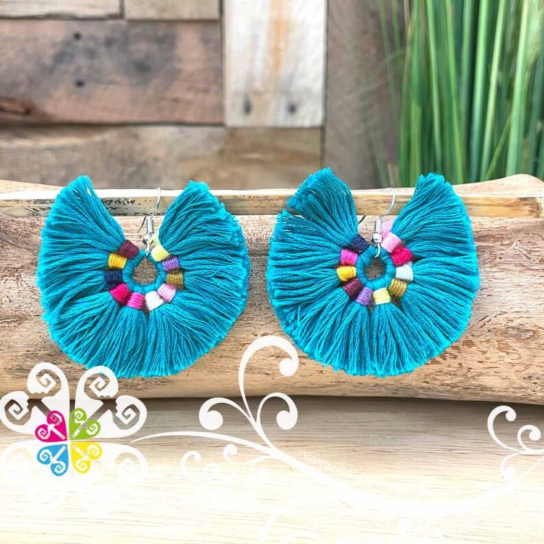 May include: A pair of turquoise tassel earrings. Each earring features a fan-shaped design with a colorful, circular accent in the center. The earrings are displayed on a wooden surface, showcasing their vibrant color and texture.