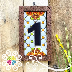 May include: A ceramic house number tile with the number "1" in black on a white background with a geometric pattern of orange, yellow, and green circles. The tile is framed in a brown wooden frame.