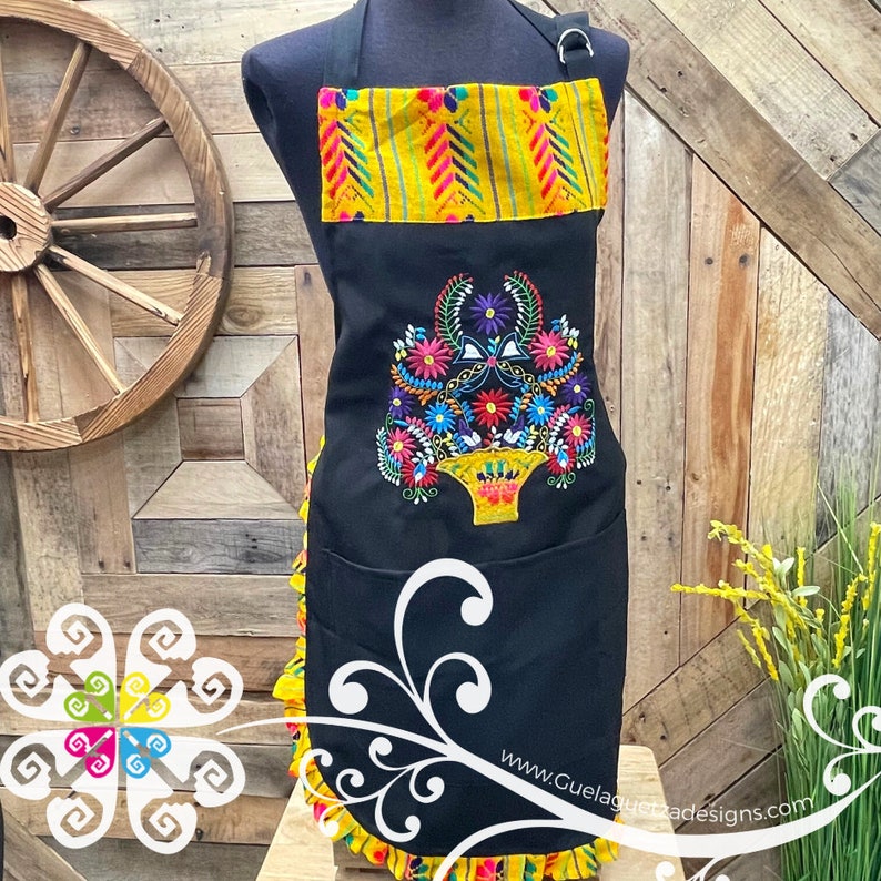 May include: Black apron with a yellow and red striped top and a colorful floral embroidery design. The apron has a large pocket and a ruffled bottom.