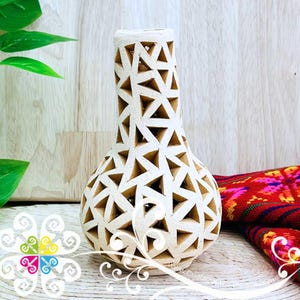May include: A cream-colored vase with a geometric triangle pattern cut out of the surface. The vase has a rounded base and a long, slender neck. The background includes a wooden surface and a colorful textile.